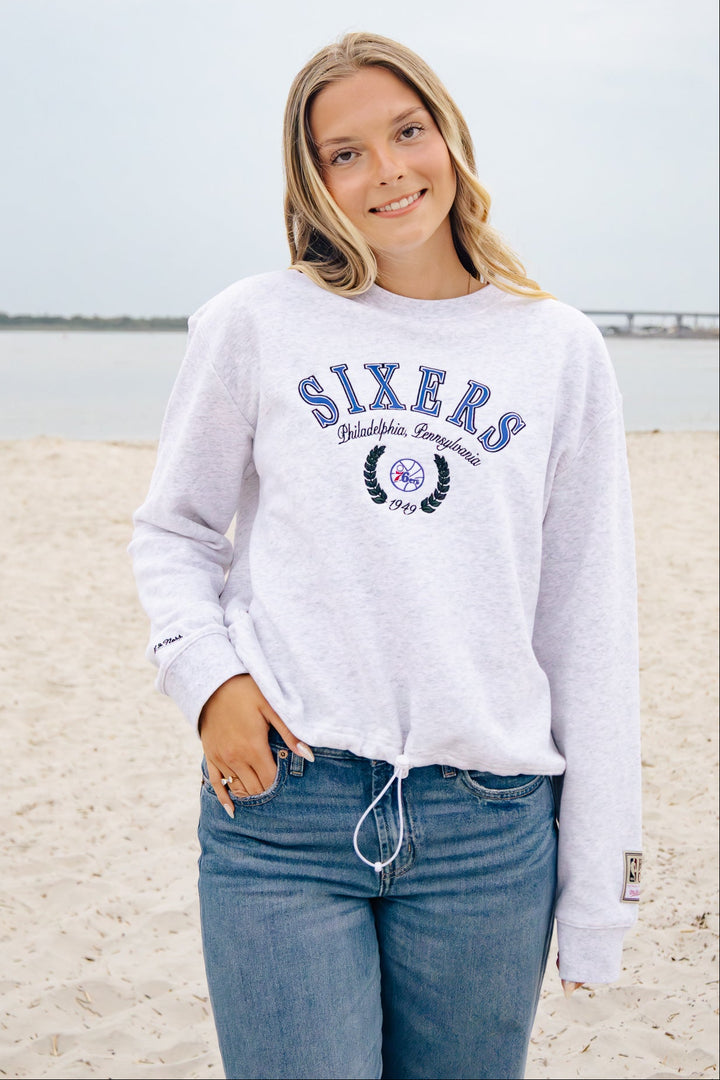 A woman standing on a sandy beach wearing a light heather gray sweatshirt with &
