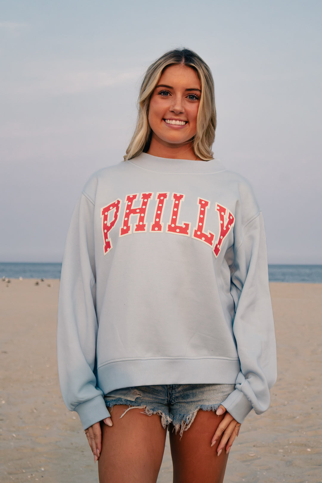 A woman standing on a beach wearing a light blue crewneck sweatshirt with 'PHILLY' printed across the chest in red varsity letters, paired with denim cutoff shorts.