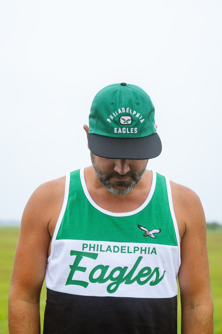 A man wearing a green and white Philadelphia Eagles baseball cap and a matching green, white, and black athletic tank top, standing outdoors against a bright sky.