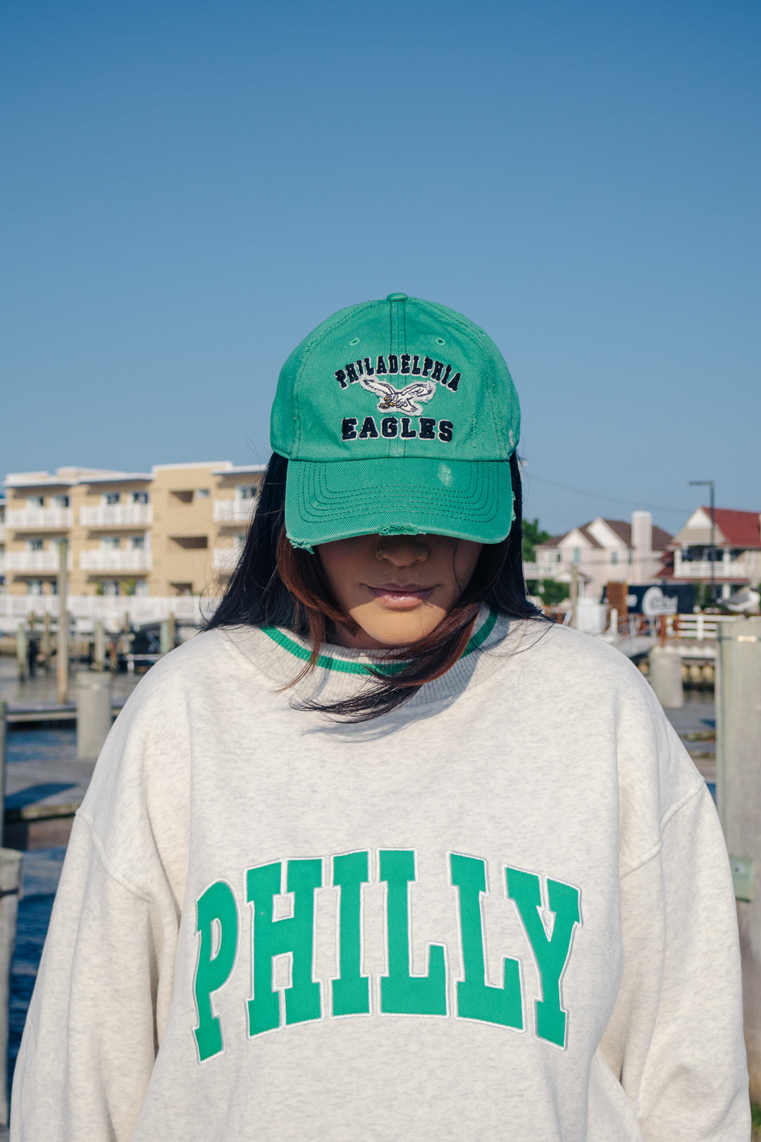 A woman outdoors wearing a kelly green Philadelphia Eagles distressed baseball cap and a light gray sweatshirt with green &