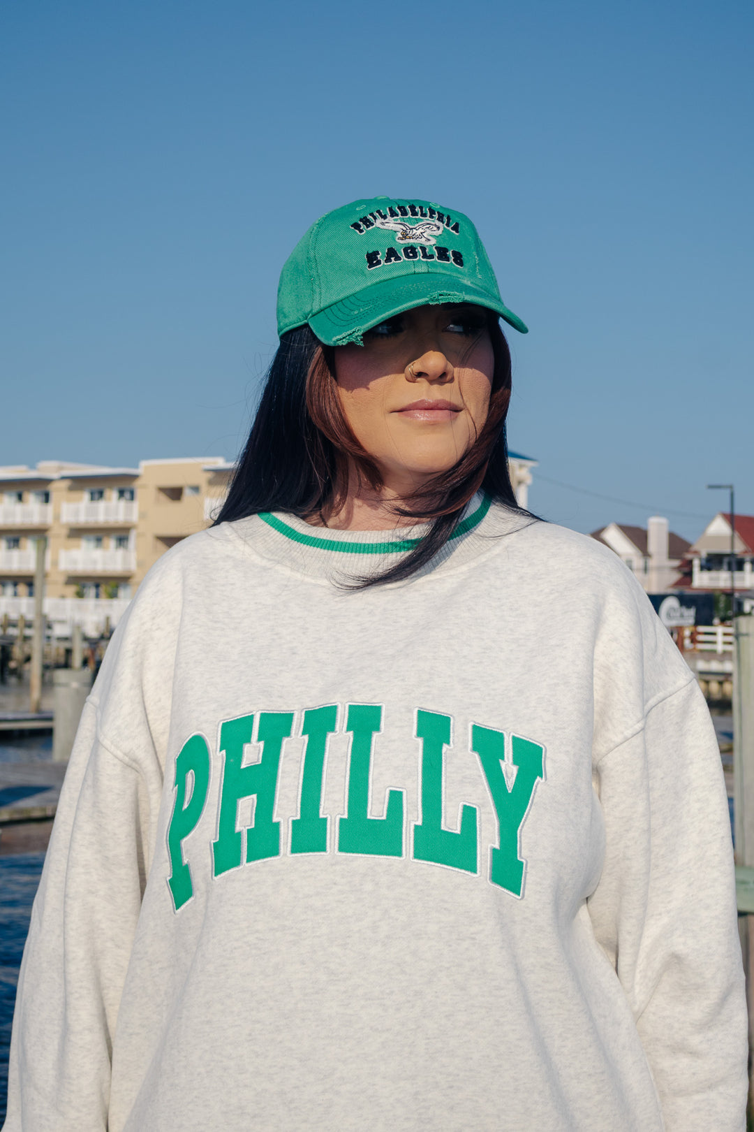 A woman outdoors wearing a kelly green Philadelphia Eagles distressed baseball cap and a light gray sweatshirt with green &
