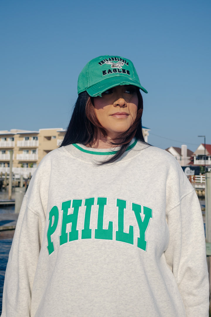 A woman outdoors wearing a kelly green Philadelphia Eagles distressed baseball cap and a light gray sweatshirt with green &