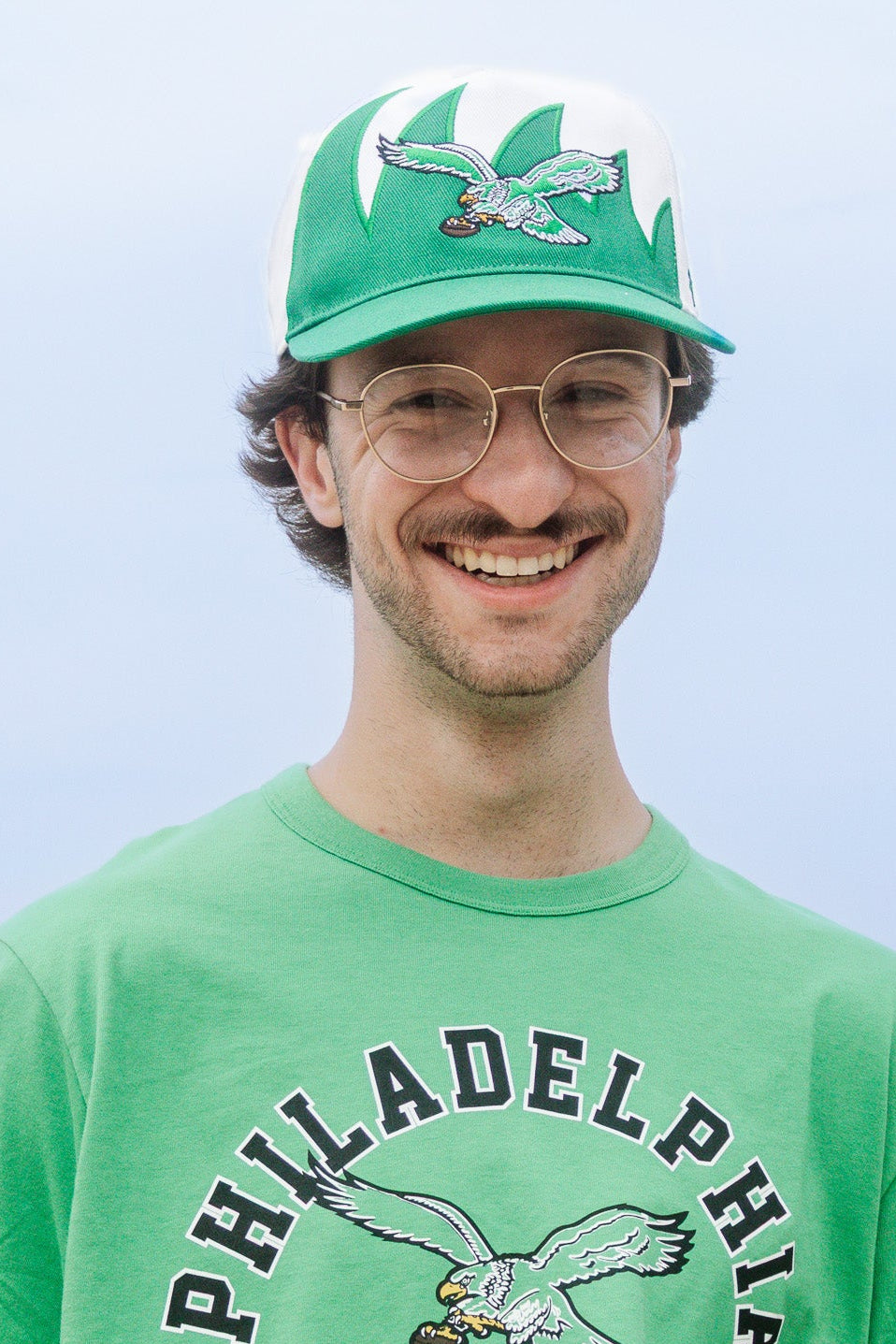 A smiling man wearing a bright green Philadelphia Eagles t-shirt and a matching green and white cap, photographed outdoors against a pale sky.
