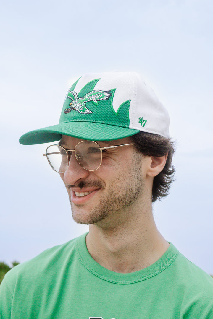 A man wearing a green and white 47 brand baseball cap with a green leaf logo, glasses, and a green t-shirt, photographed outdoors against a pale sky.