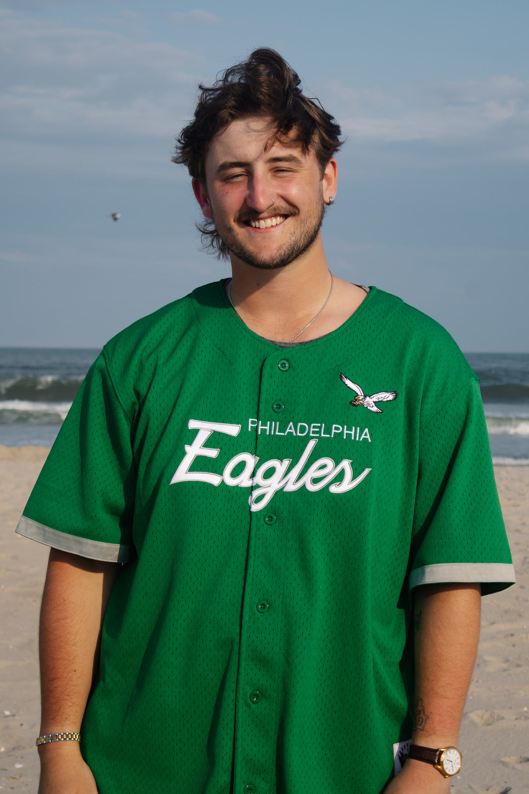 A smiling man standing on a beach wearing a green Philadelphia Eagles button-up baseball jersey with white lettering and gray sleeve trim.