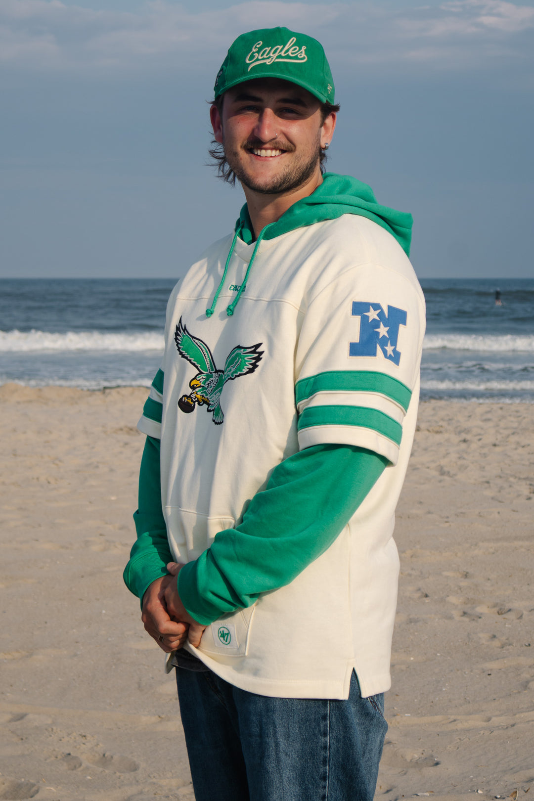 A man standing on a sandy beach wearing a green and off-white pullover hoodie with a large old Eagles logo graphic on the front.