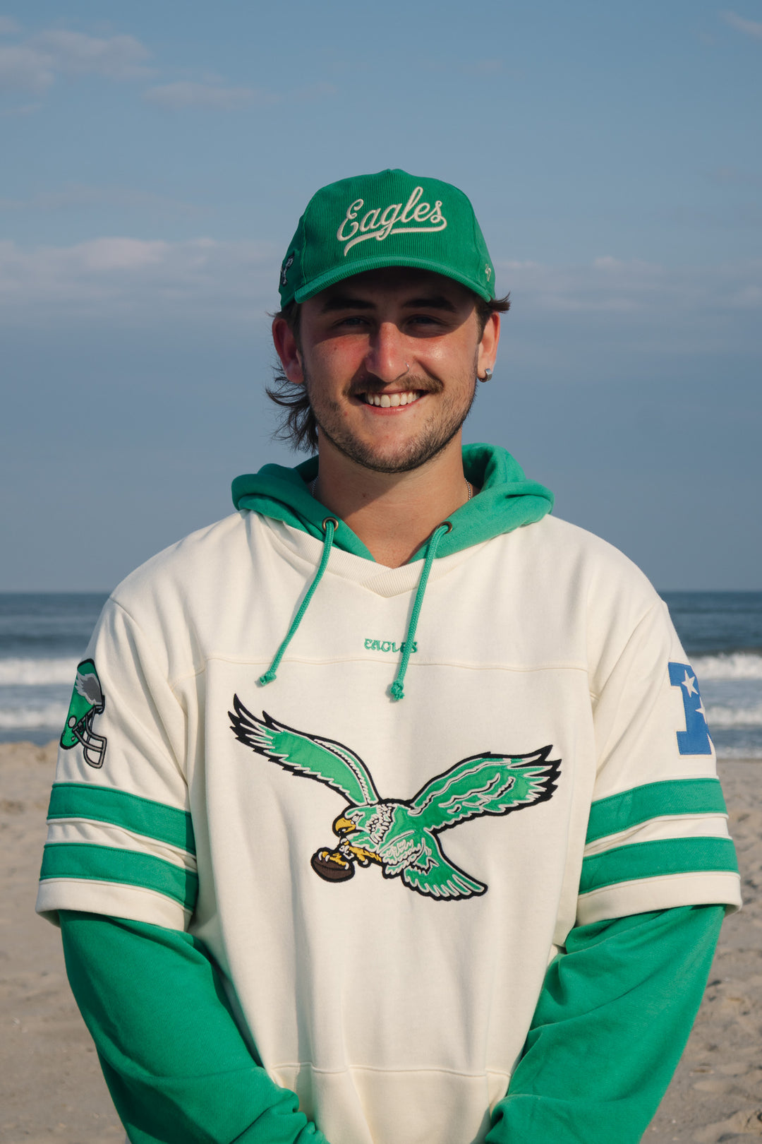 A smiling man standing on a beach wearing a green and white Eagles hoodie with a large eagle graphic on the front, a green Philadelphia Eagles corduroy adjustable cap, and the ocean and sky in the background.