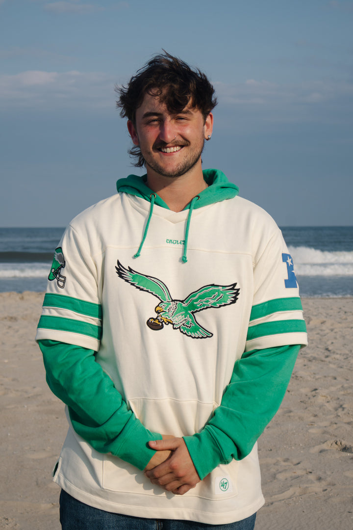 A man standing on a sandy beach wearing a green and off-white pullover hoodie with a large old Eagles logo graphic on the front.