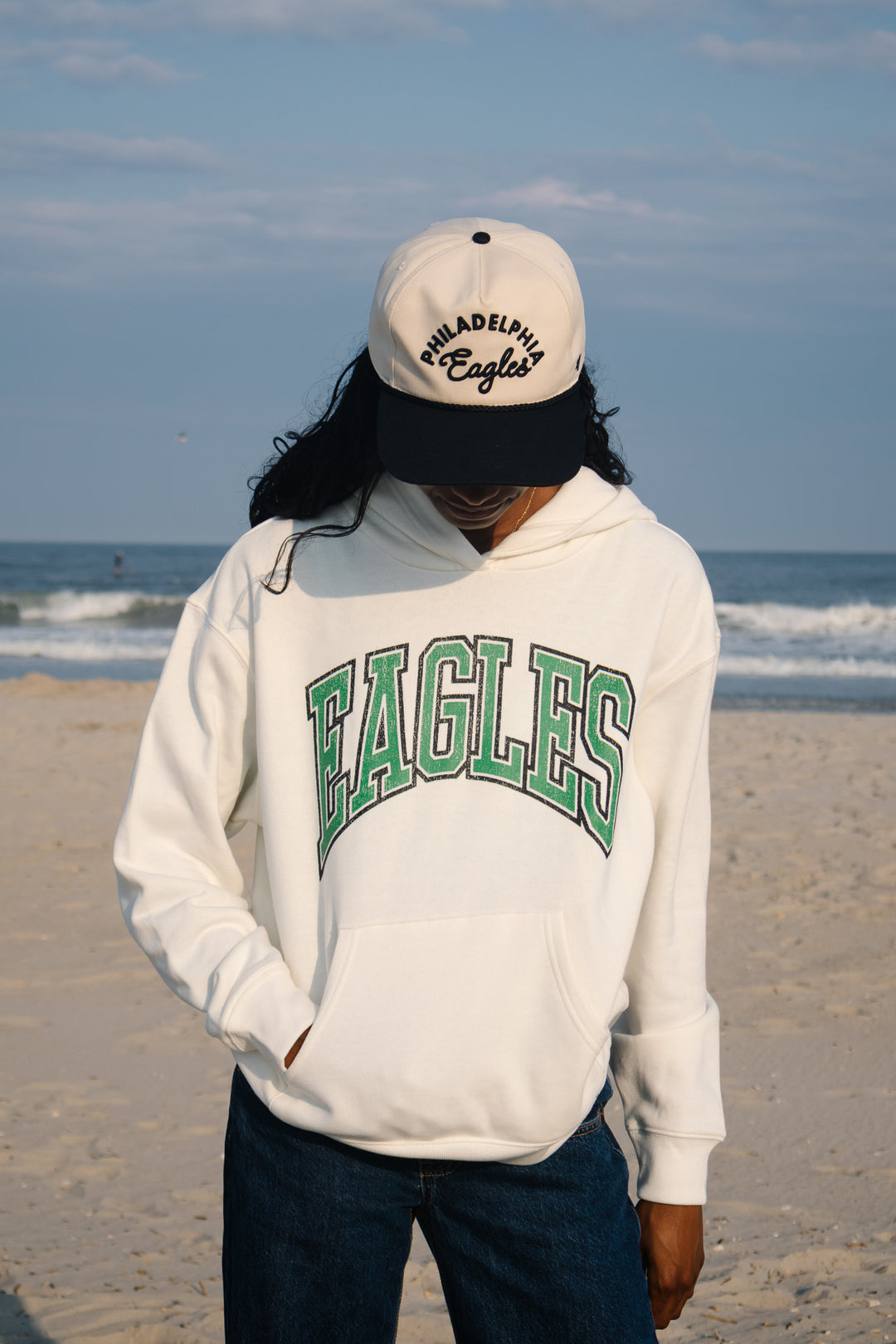 A person standing on a sandy beach wearing a cream hoodie with green and black &