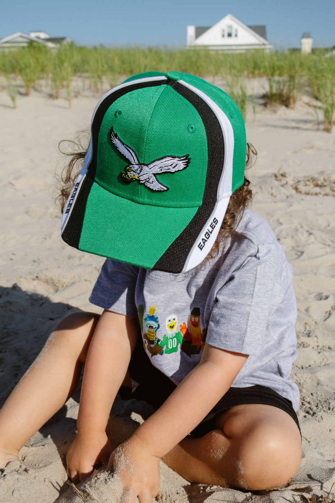 A child sitting on a sandy beach wearing a green Philadelphia Eagles snapback adjustable hat and a gray t-shirt.