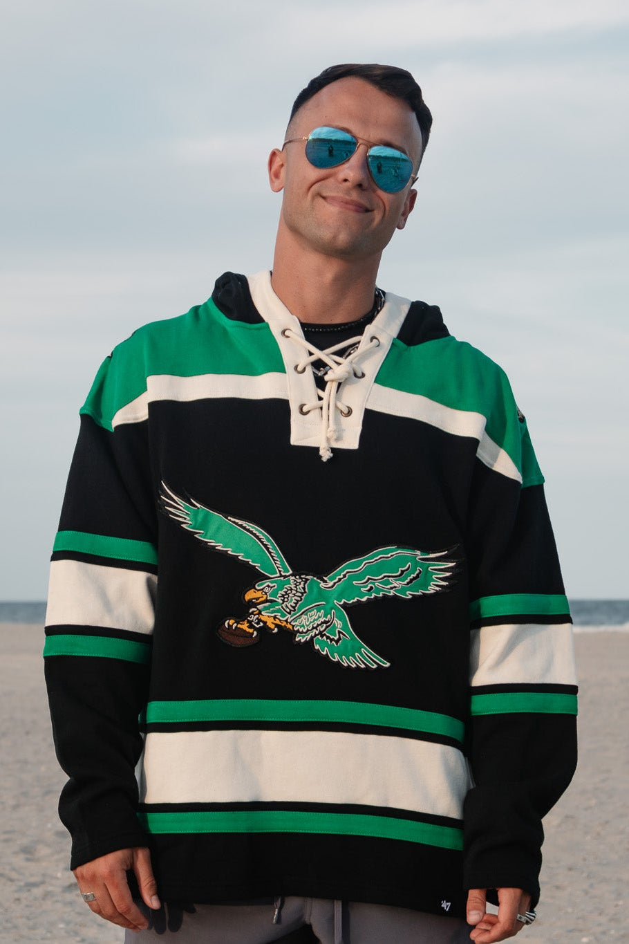 A man wearing a green, black, and white hockey-style hoodie with a large old school Eagles graphic/logo on the front, standing on a sandy beach with the ocean and cloudy sky in the background.