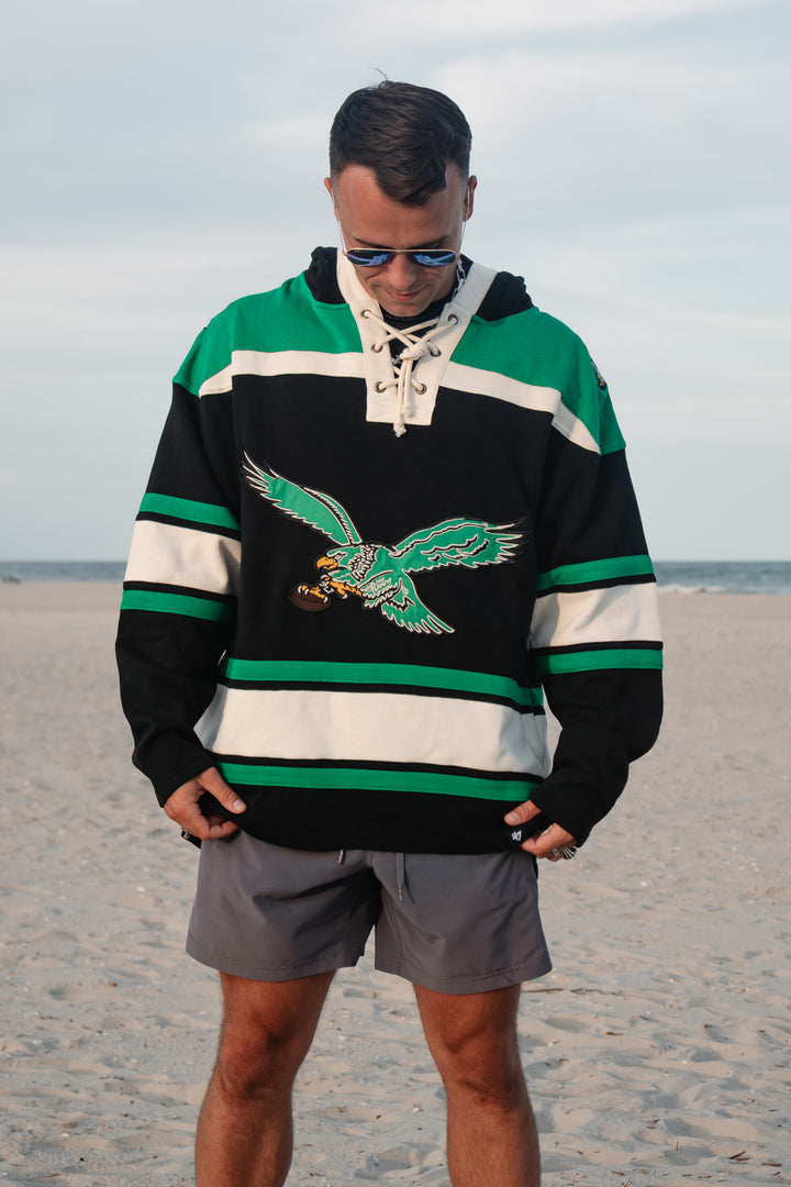 A man wearing a green, black, and white hockey-style hoodie with a large old school Eagles graphic/logo on the front, standing on a sandy beach with the ocean and cloudy sky in the background.