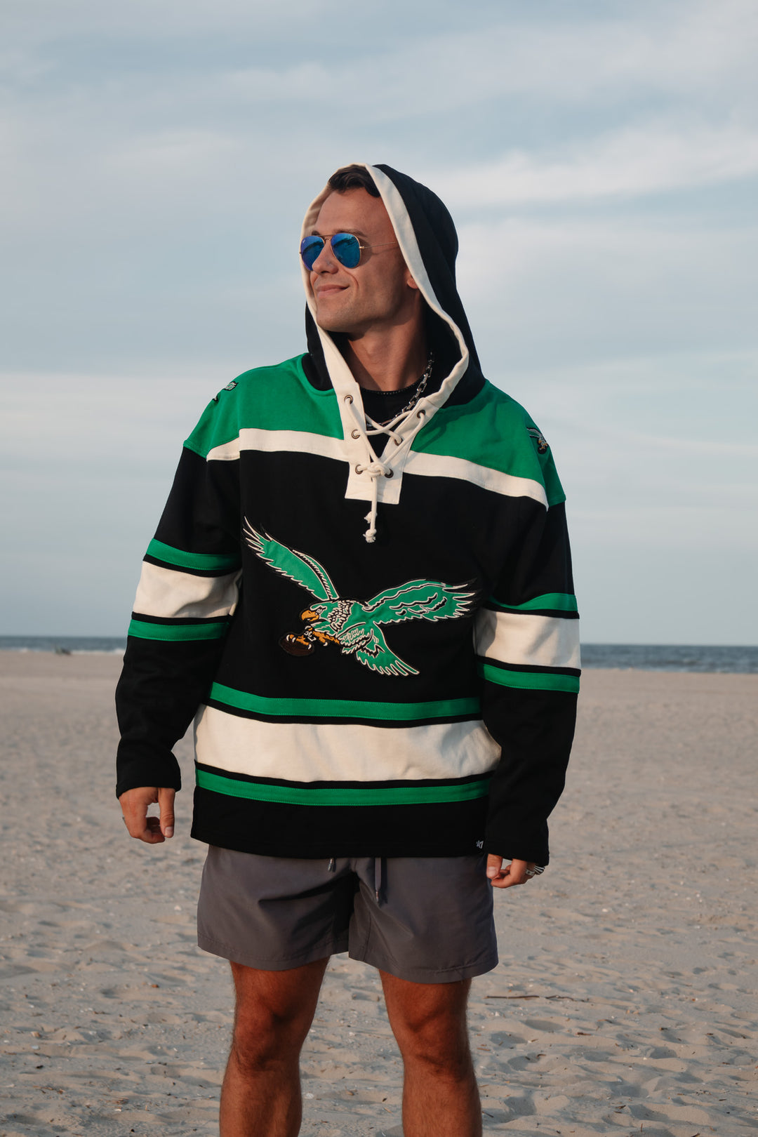 A man wearing a green, black, and white hockey-style hoodie with a large old school Eagles graphic/logo on the front, standing on a sandy beach with the ocean and cloudy sky in the background.