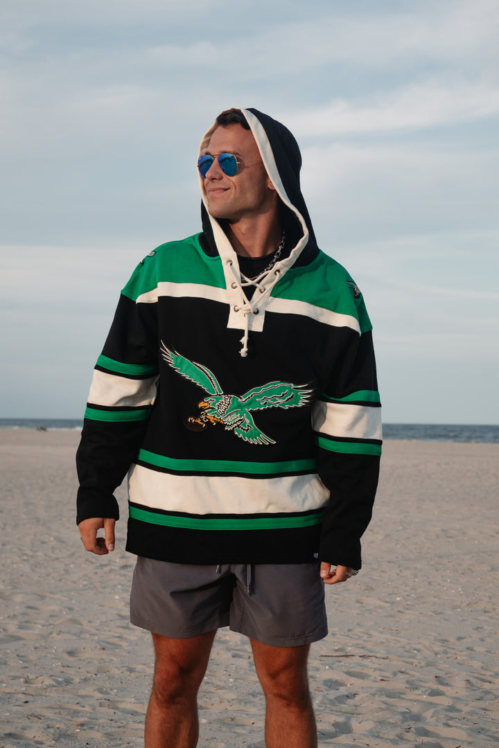 A man wearing a green, black, and white hockey-style hoodie with a large old school Eagles graphic/logo on the front, standing on a sandy beach with the ocean and cloudy sky in the background.